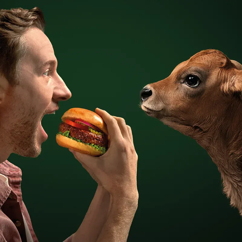 Social Ad von Beyond Meat im Format 1:1 aus der Kampagne All Taste No Worries: ein Mann beißt in einen Beyond Burger, ihm gegenüber steht ein Kalb vor dunkelgrünem Hintergrund